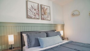 A neatly made bed with a blue comforter and pillows, flanked by bedside tables with lamps, in a room with a green paneled headboard and two framed floral artworks on the wall.