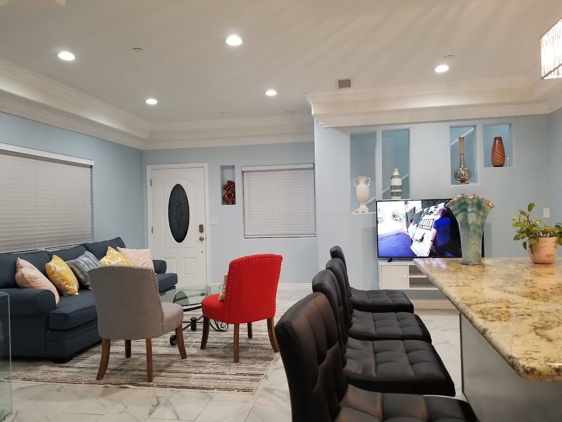 Un salon moderne avec un canapé bleu, un fauteuil rouge et un pouf noir. Un téléviseur à écran plat est fixé au mur, et des objets décoratifs sont disposés sur les étagères.