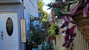 Un estrecho callejón con una puerta blanca y un letrero que dice 'WELCOME', flanqueado por macetas con plantas y un edificio azul.