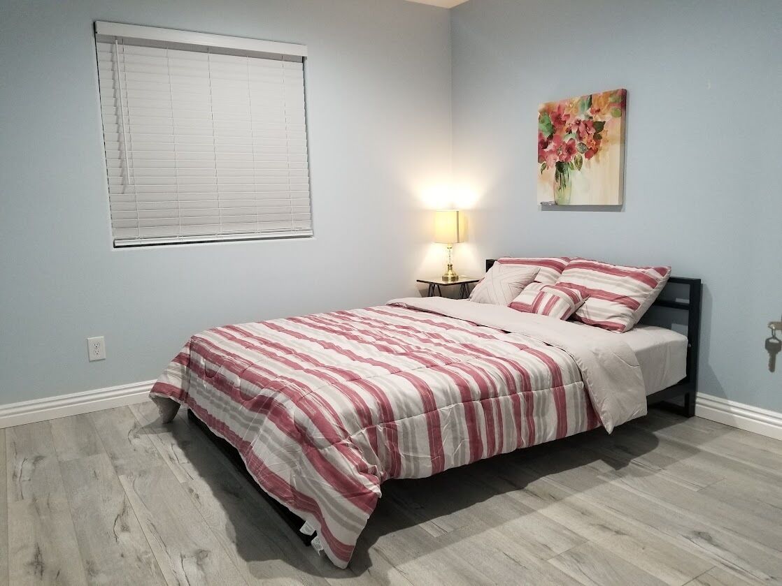 A bedroom with a bed, a nightstand, a lamp, a window with blinds, and a painting on the wall.