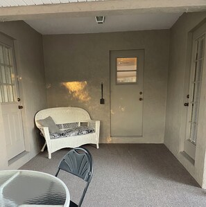 A covered porch with a wicker chair and a small table, a door with a window, and a textured wall. - Pinecraft - the House at Phillippi Crossing - 3 bedroom, 3 bathroom (Sarasota)