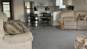 En moderne stue med sofa, to lænestole, sofabord og spiseplads med et køkken i baggrunden.