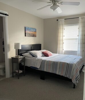 A bedroom with a bed, a nightstand, a ceiling fan, a window with curtains, and a painting on the wall. - Pinecraft - the House at Phillippi Crossing - 3 bedroom, 3 bathroom (Sarasota)