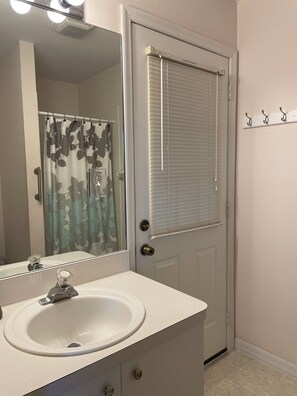 A bathroom with a white sink, a mirror, a shower curtain with a leaf pattern, and a door with a window blind. - Pinecraft - the House at Phillippi Crossing - 3 bedroom, 3 bathroom (Sarasota)