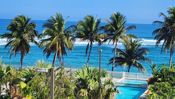A pool area with a view of the ocean and palm trees. - Ocean Front 3-bedroom condo with Pool, AC, WiFi, 2 King, 1 Queen. (AGUADA)