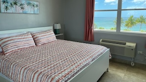 A bedroom with a bed, a nightstand, a lamp, a window with a view of the ocean, and a painting on the wall. - Ocean Front 3-bedroom condo with Pool, AC, WiFi, 2 King, 1 Queen. (AGUADA)