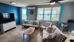 A living room with a flat-screen TV, two sofas, a coffee table, and a desk with a chair. - Ocean Front 3-bedroom condo with Pool, AC, WiFi, 2 King, 1 Queen. (AGUADA)