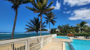 A pool area with a view of the ocean, surrounded by palm trees and a white fence. - Ocean Front 3-bedroom condo with Pool, AC, WiFi, 2 King, 1 Queen. (AGUADA)