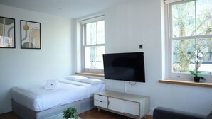 A modern hotel room with a bed, a flat-screen TV mounted on the wall, a small bedside table with a plant, and a window with a view of trees.