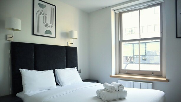 A neatly made bed with white linens and a rolled towel on the side, a black headboard, and a window with a view of the city.