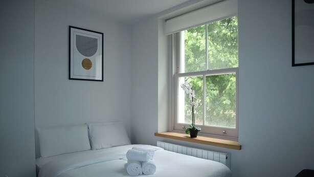 A neatly made bed with white linens, a framed artwork above it, and a window with a view of trees.