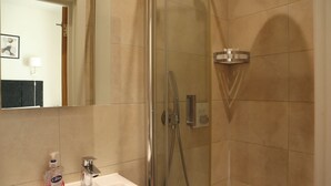 Un baño con ducha envuelta en vidrio, un lavamanos con jabón para manos y un espejo que refleja una puerta y un aplique en la pared.