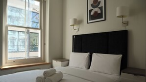 Una habitación de hotel con una cama grande, dos mesitas de noche, un ventanal con vistas a edificios y una lámpara de pared.