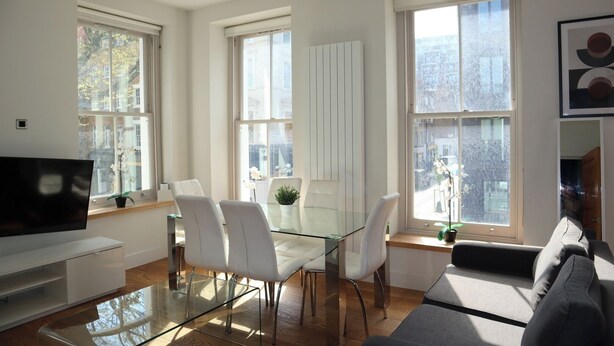 Una sala moderna con una mesa de comedor de vidrio, sillas blancas, un televisor de pantalla plana y ventanas amplias que ofrecen vistas de la ciudad.