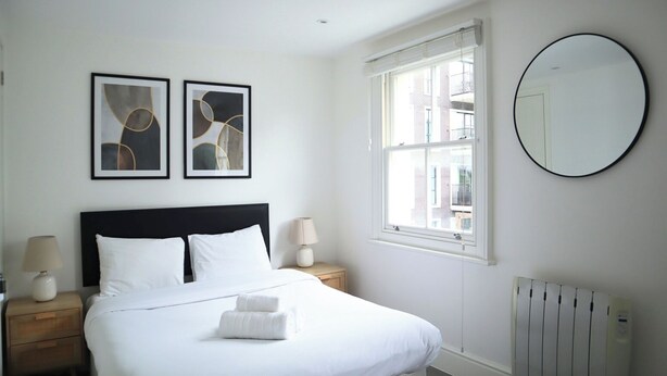 A neatly arranged bedroom with a large bed, two bedside tables with lamps, a window with blinds, and two framed abstract artworks on the wall.