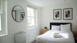 A bedroom with a bed, a round mirror, two framed artworks, a radiator, and a window.