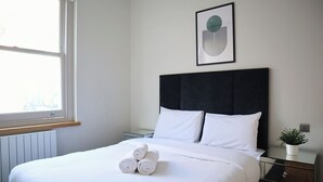 Ein ordentlich bezogenes Bett mit weißen Leinen und aufgerollten Handtüchern, ein schwarzer möbelbezogener Kopfteil, ein gerahmtes abstraktes Kunstwerk, eine Topfpflanze und ein Fenster mit Blick auf die Stadt.