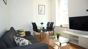 Ein modernes Wohnzimmer mit einem Glastisch, einer Couch mit gemusterten Kissen, einem Essbereich mit einem Glastisch und schwarzen Stühlen sowie einem flachbildfernseher an der Wand.