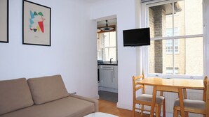 A compact living space with a sofa, a dining table with chairs, a TV mounted above the table, and a window with a view of the outside.