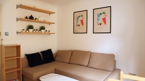 A living room with a beige sectional sofa, a wooden side table, and a shelf with potted plants.