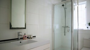 Un baño moderno con ducha de vidrio, lavamanos blanco con espejo y artículos de aseo.