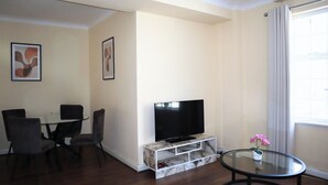 Una sala de estar con un televisor sobre un pedestal de mármol, una mesa de comedor con sillas y una mesita con una planta en maceta.