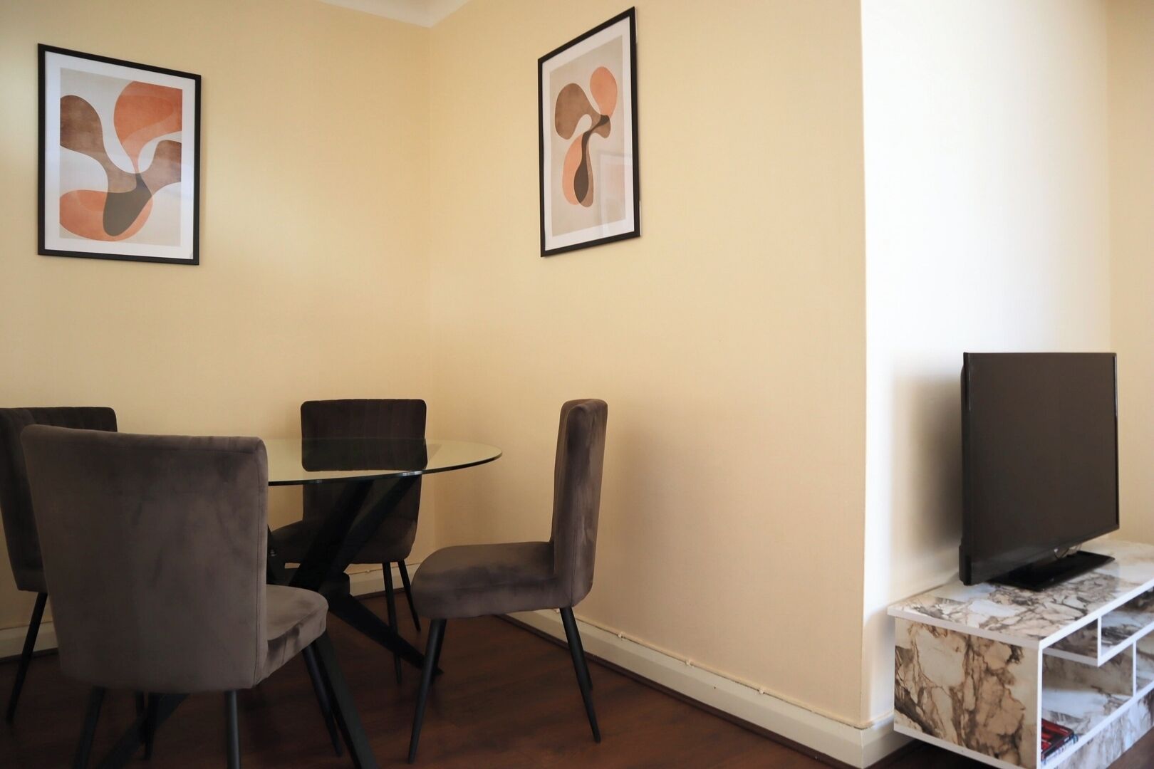 A dining area with a glass table and four chairs, a flat-screen TV on a marble console, and abstract art on the walls.