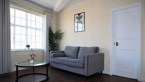 Un salón con un sofá gris, una mesa redonda de vidrio con una orquídea en maceta, un ventanal con cortinas, una planta en maceta y una obra de arte abstracto enmarcada en la pared.