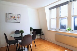 Uma sala de estar moderna com uma mesa de vidro, cadeiras escuras, uma televisão de tela plana e uma janela grande com plantas.