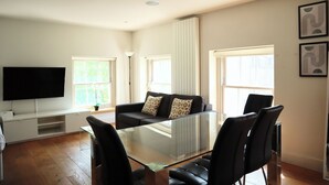 Een moderne woonkamer met een glazen eettafel, zwarte stoelen, een zwarte bank met gekleurde kussens, een flatscreen televisie en grote ramen met witte gordijnen.