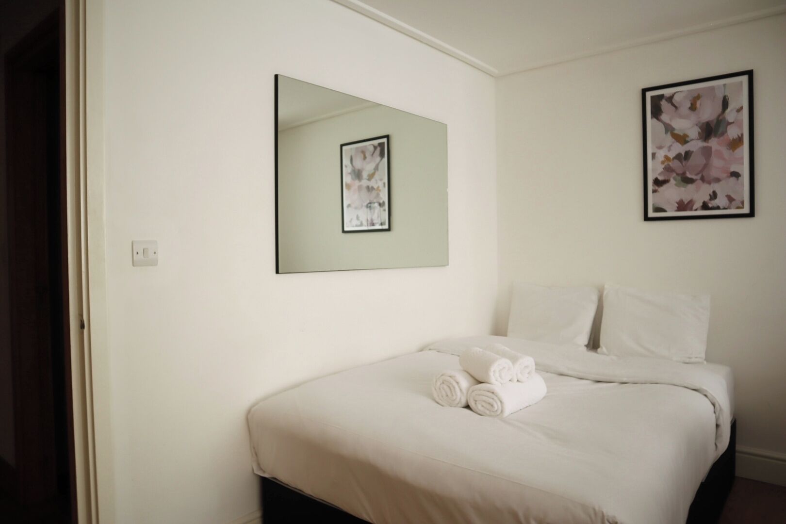 A neatly made bed with white linens and rolled towels, a large mirror on the wall, and a framed floral artwork above the bed.