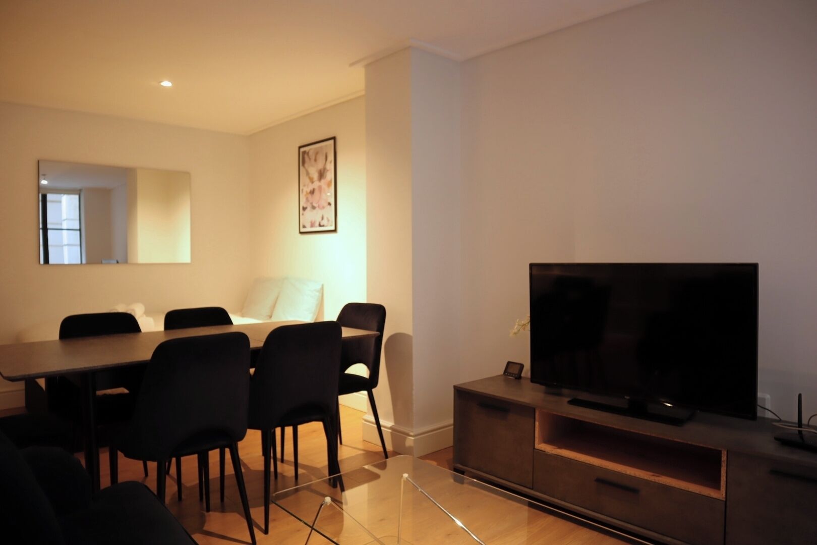 Una sala de estar moderna con un televisor de pantalla plana sobre un soporte de madera, un comedor con mesa y sillas, y un espejo grande en la pared.