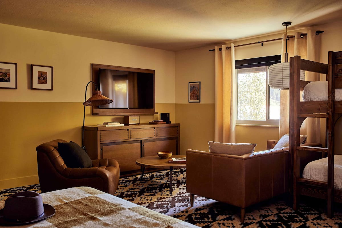 A cozy room with a sofa, armchair, and a bunk bed. There is a television on the wall, a lamp, and a window with curtains.