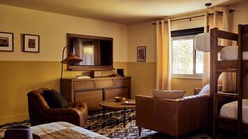 A cozy room with a sofa, armchair, and a bunk bed. There is a television on the wall, a lamp, and a window with curtains.