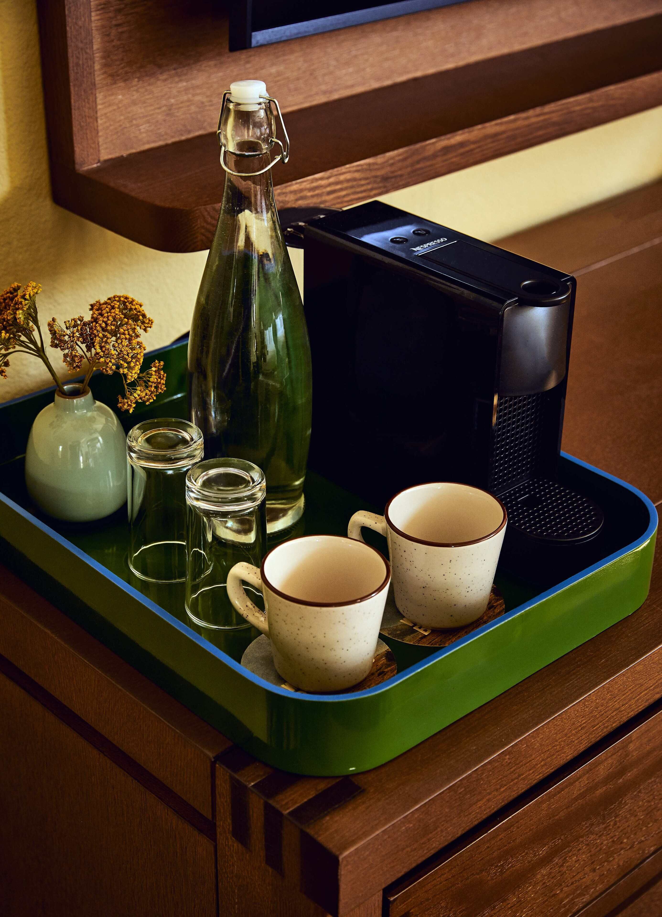 A tray with a green tray, a glass bottle, two mugs, a small vase with dried flowers, and a water glass.