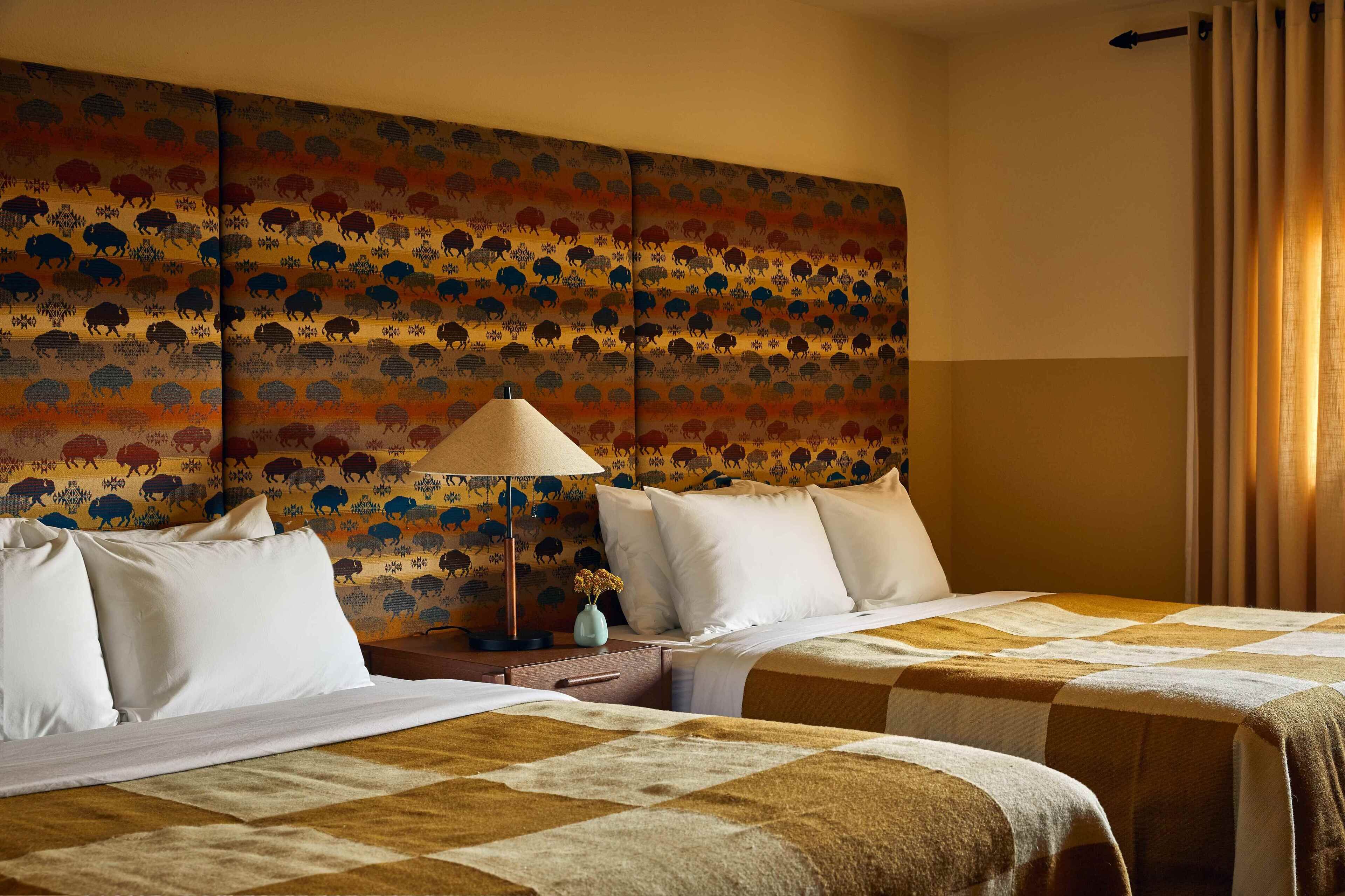 Una habitación de hotel con dos camas, un cabecero estampado, una mesita de noche con lámpara y una ventana con cortinas.