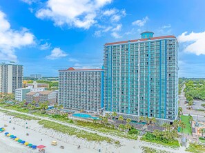A high-rise hotel with multiple balconies, a swimming pool, and a beach view.