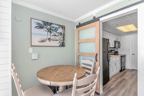 A dining area with a round wooden table, white chairs, a beach-themed painting on the wall, and a kitchen visible through an open door.