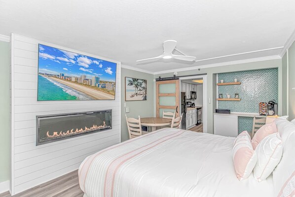 A bedroom with a bed, a fireplace, a ceiling fan, a kitchen area, and a beach view.