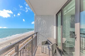 A balcony with a wicker chair, a glass door, and a view of the beach and ocean. - Oceanfront Luxe 1BR w/ Pools & Waterpark! (Myrtle Beach)