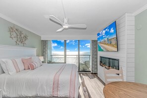 A bedroom with a large bed, a ceiling fan, a TV, a fireplace, and a balcony with a city view.