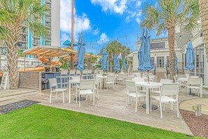 An outdoor patio with white tables and blue umbrellas, surrounded by palm trees and a building with balconies.