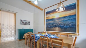 Un coin repas avec une nappe à carreaux, des chaises en bois, un buffet de couleur turquoise et un grand tableau représentant un coucher de soleil sur l’océan.