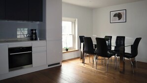 A modern kitchen with white cabinets, a built-in oven, and a coffee maker. There is a dining area with a glass table and black chairs. A window provides natural light.