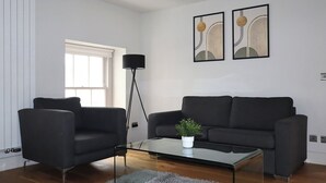 A modern living room with a black sofa, two armchairs, a glass coffee table with a plant, and abstract wall art.