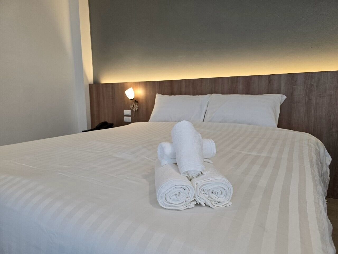 A neatly made bed with white linens and rolled towels, a wooden headboard, and a bedside lamp.