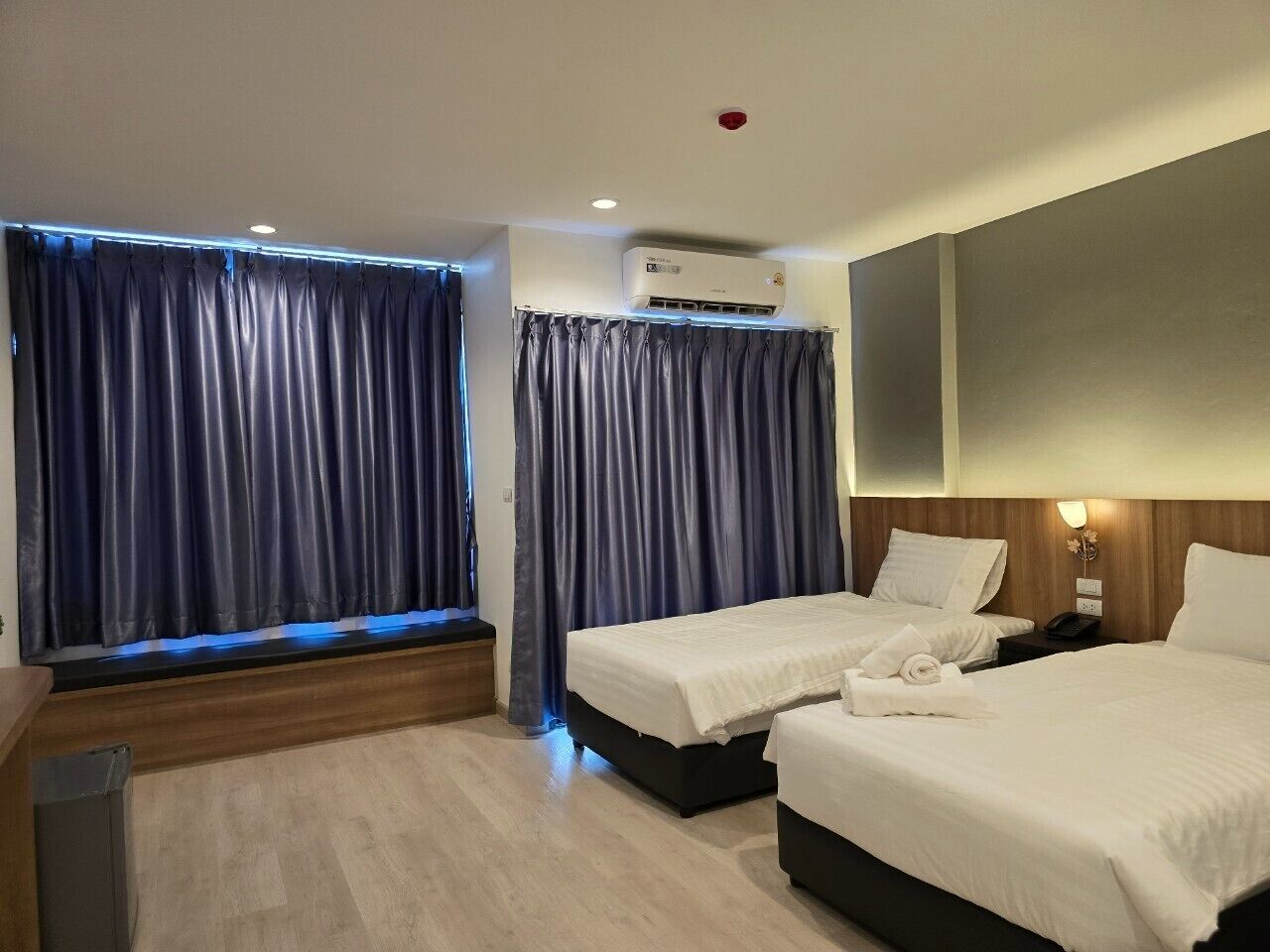Habitación de hotel con dos camas, una ventana con cortinas, un aire acondicionado y una mesita de noche con lámpara.