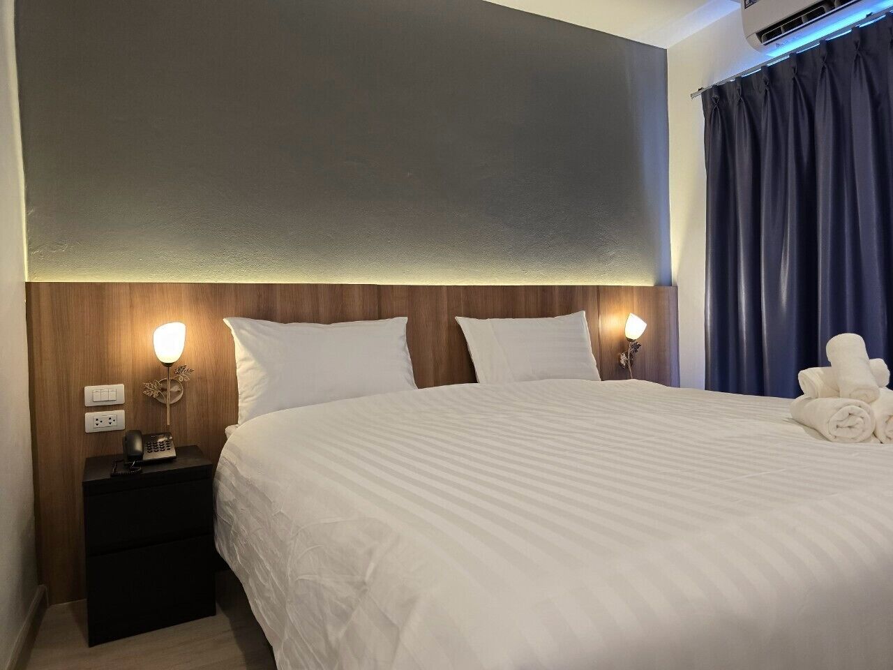 A hotel room with a large bed, two bedside tables with lamps, a wooden headboard, and a dark curtain.