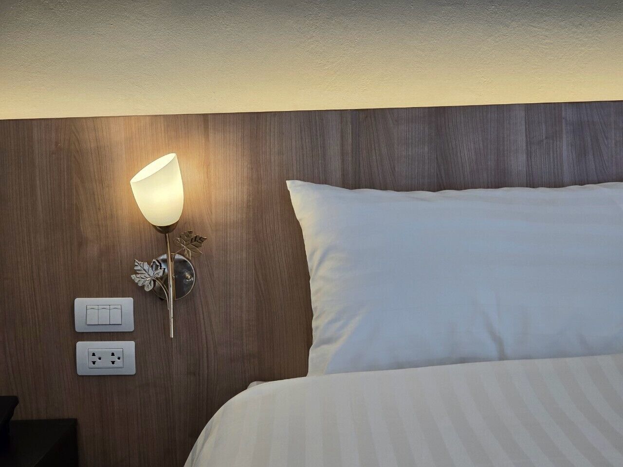 A hotel room with a wooden headboard, a bedside lamp, a power outlet, and a bed with white bedding.