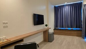 A modern hotel room with a flat-screen TV mounted on the wall, a wooden bench, a small refrigerator, and a window with dark curtains.